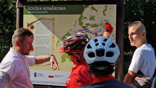 Uczestnicy otwarcia stoją czy tablicy informacyjnej, pokazują szlak jakim prowadzi ścieżka rowerowa. 