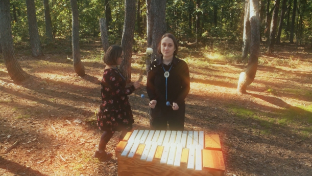 kadr z teledysku, dwie aktorki w krzywm lesie pod Gryfinem, jedna tańczy, druga gra na cymbałach 
