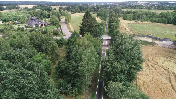 Sfotografowany z lotu ptaka długi prosty odcinek drogi rowerowej o nawierzchni bitumicznej. Nad nią jest mały wiadukt umożliwijący przejazd samochodem. Po obu stronach trasy rowerowej - drzewa, łąki i pola. Po lewej - duży dom. W oddali - miasteczko.